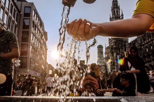 Klimawandel und Hitze in der Stadt Blick in eine Innenstadt mit Häusern und Menschen, im Vordergrund eine Hand, die Wasser aus einem Hahn oder Brunnen schöpft.